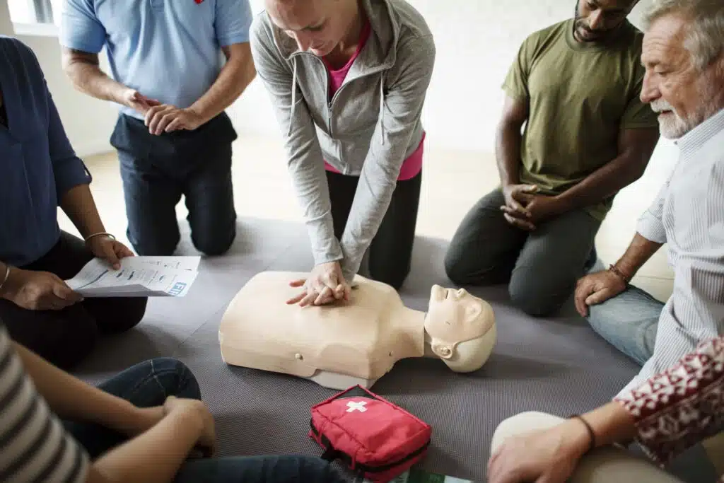 A group of people watch as one person practises chest compressions on a CPR training manikin, with a first aid kit and instruction sheet nearby.