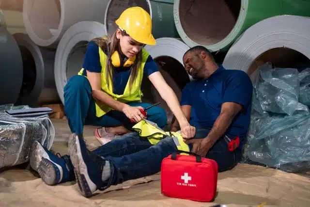 A construction worker in a safety vest and helmet applies a tourniquet to an injured man's leg while he sits in pain on the floor. A red first aid kit is open nearby. Large industrial pipes are in the background.