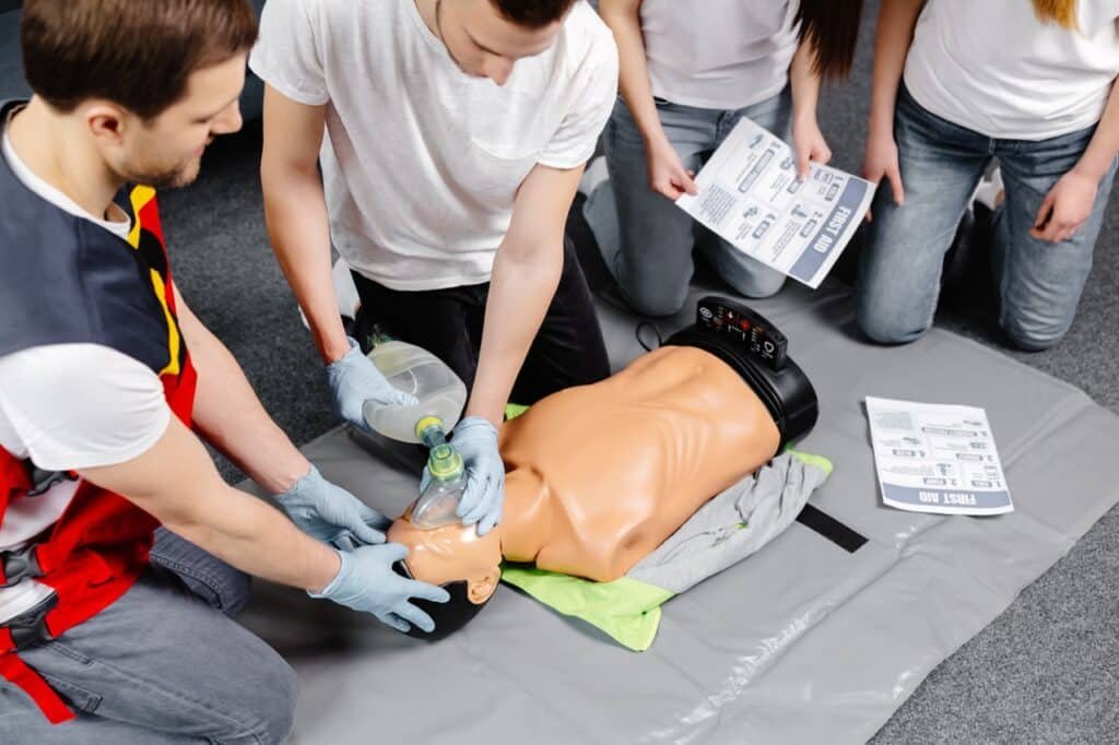 Four people practise CPR on a medical dummy, with two performing resuscitation using a bag valve mask and two holding instruction sheets. The group is kneeling on a mat, focused on learning emergency procedures.