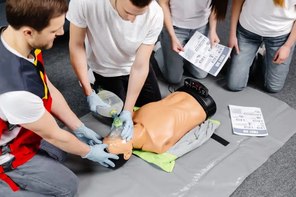 Four people practise CPR on a medical dummy, with two performing resuscitation using a bag valve mask and two holding instruction sheets. The group is kneeling on a mat, focused on learning emergency procedures.