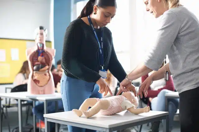 Two women practise infant CPR on a baby manikin on a table in a classroom, with a human anatomy model and other people visible in the background.