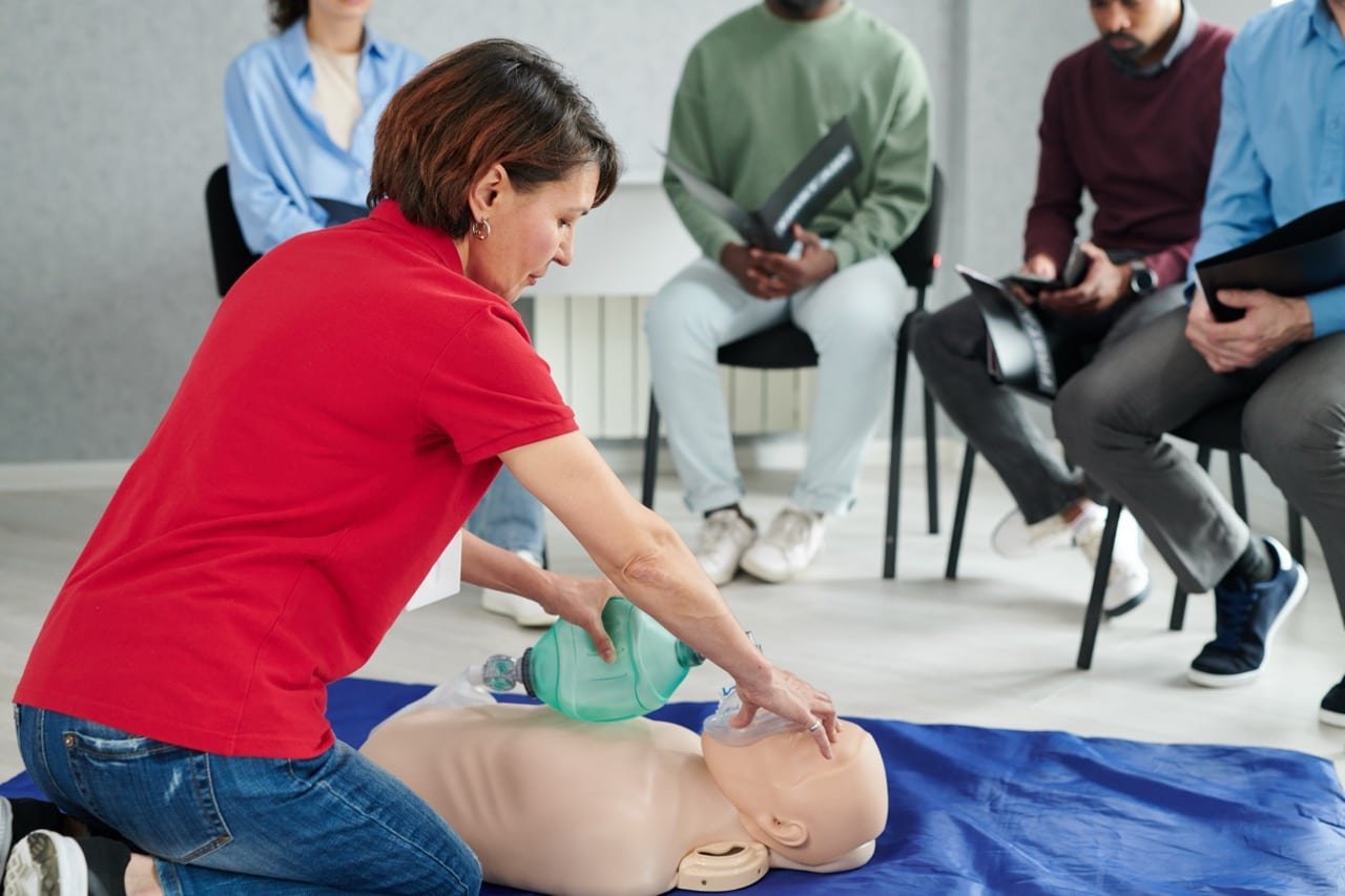 A woman in a red shirt demonstrates CPR with a resuscitation bag on a practice dummy whilst several people sit in chairs watching and taking notes in a classroom setting.