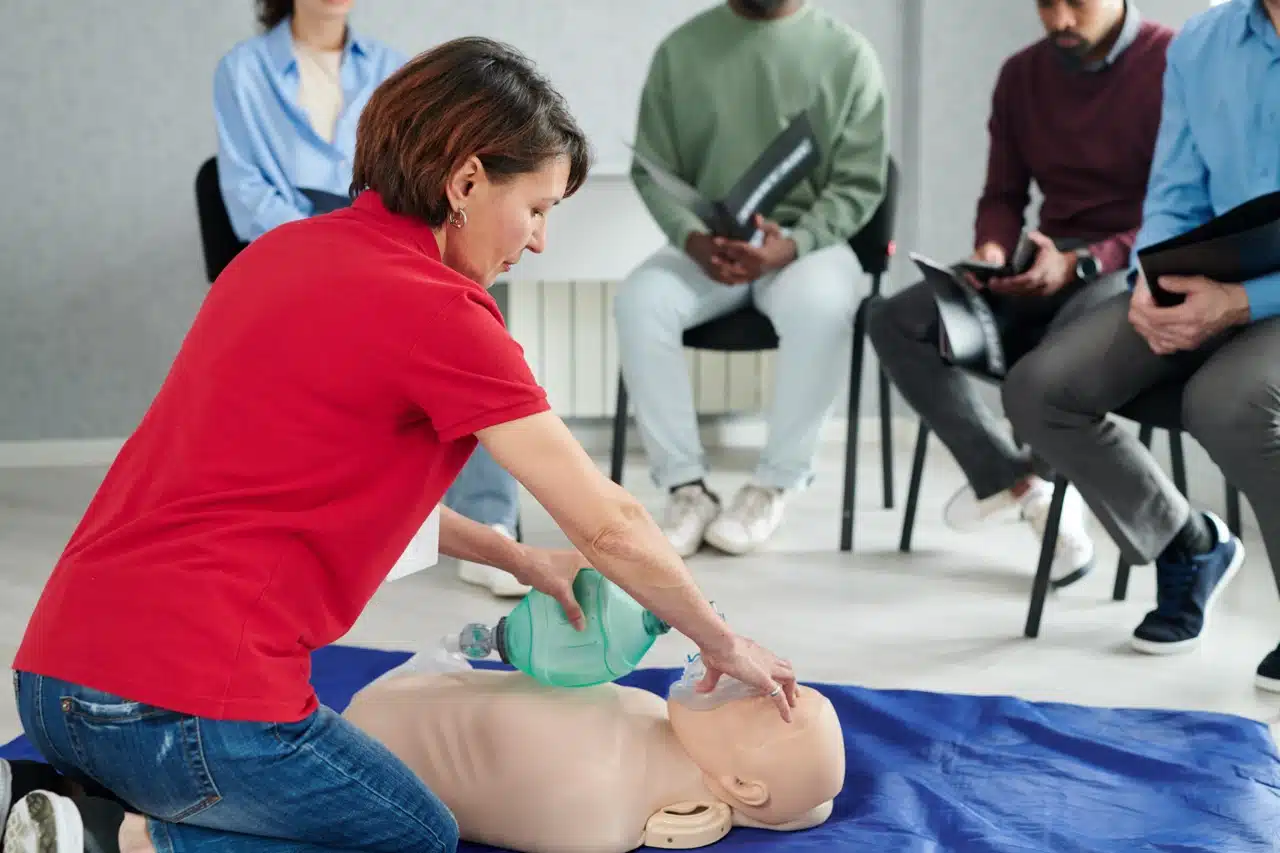 A woman in a red shirt demonstrates CPR with a resuscitation bag on a practice dummy whilst several people sit in chairs watching and taking notes in a classroom setting.